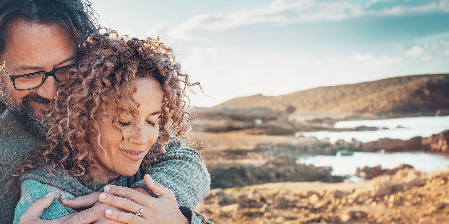 An older couple embracing each other while standing by a scenic coastal landscape with a serene expression on their faces."