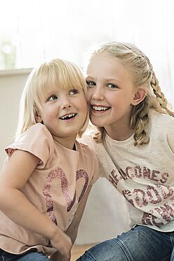 Two young girls, one blonde and the other with braided hair, sit close together, smiling and appearing happy.