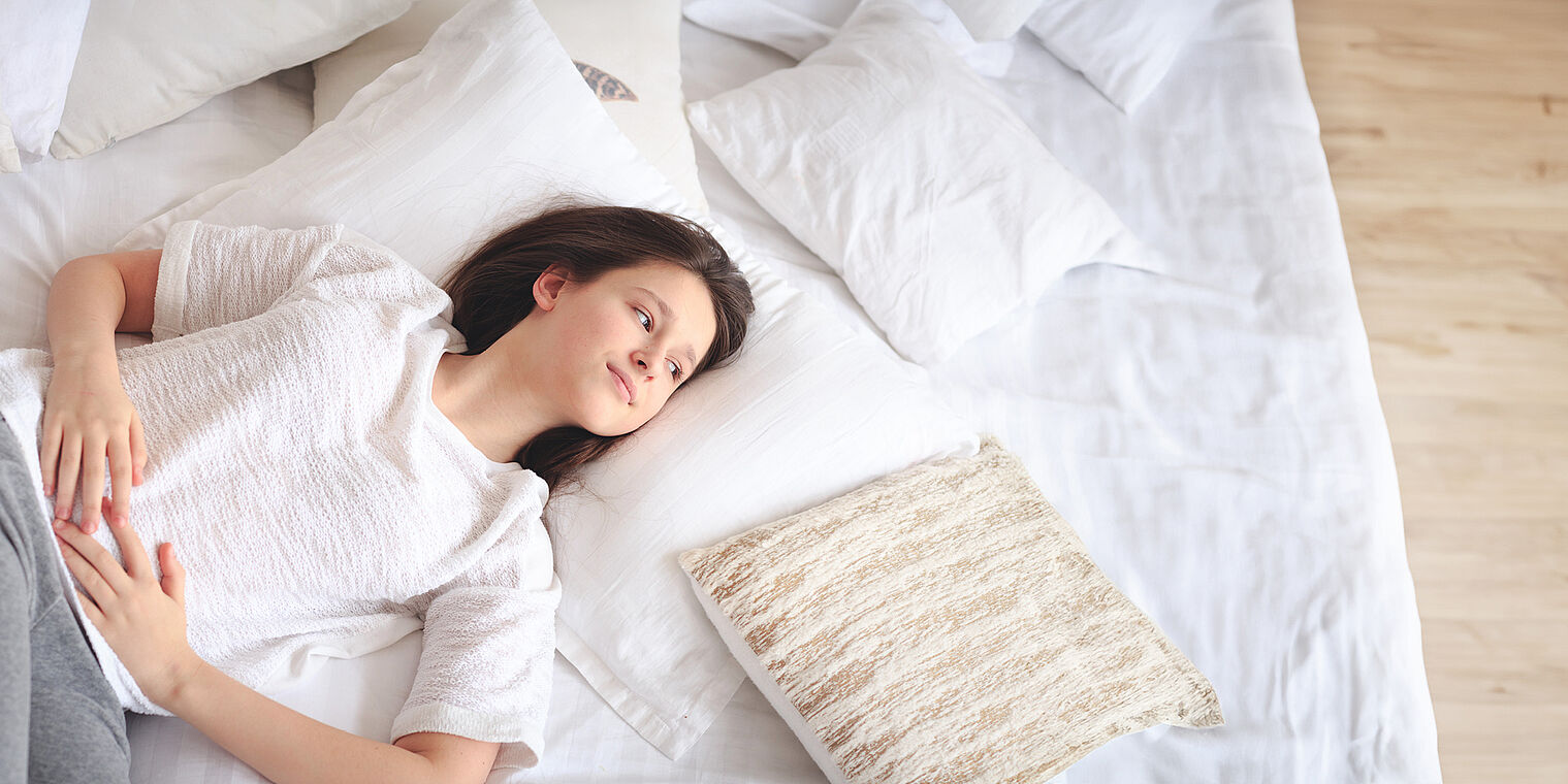 young girl lying on a bed with white sheets and pillows, looking contemplatively upwards.