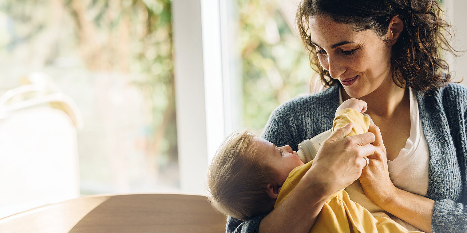 Mother feeding her baby with a bottle while lovingly looking at the child in a brightly lit room with a large window in the background