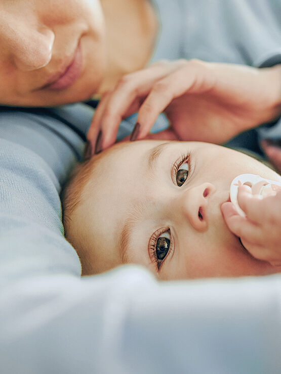 Close-up of a baby lying down, holding a pacifier, with a parent gently touching the baby's head.