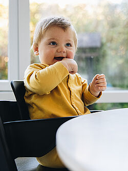 Smiling baby in a yellow outfit sitting in a high chair, looking content while putting their hand in their mouth, with a window and greenery in the background.